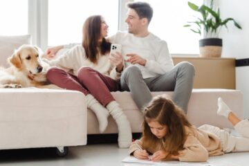 Two adults are sitting on an L-shaped sofa beside a dog. A young girl lies on her belly on the floor while drawing.
