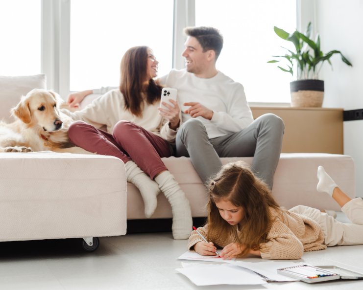 Two adults are sitting on an L-shaped sofa beside a dog. A young girl lies on her belly on the floor while drawing.