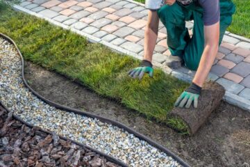 A gardener applying a line of turf onto a open patch of dirt. The dirt is next to a garden bed of rocks.