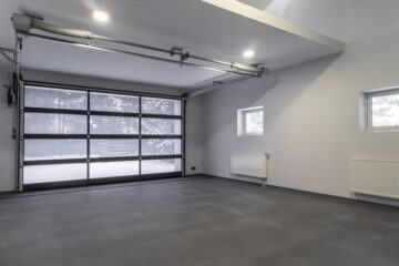 An interior view of a modern home garage with a large glass door, a grey floor, and white-painted walls.