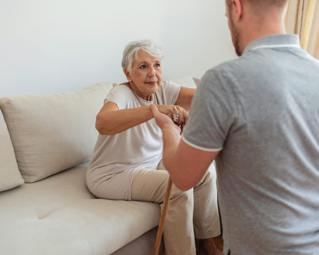 A man wearing a grey polo, kneeling and holding the hands of a senior woman sitting on a couch with a cane.