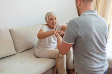 A man wearing a grey polo, kneeling and holding the hands of a senior woman sitting on a couch with a cane.