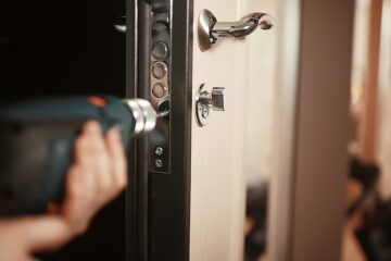 Close-up of a person's hand holding a power drill as they install a new lock on their door. The door handle is silver.