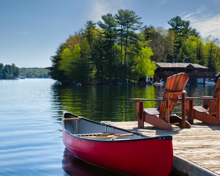 Two chairs on a wooden dock that looks out over a peaceful lake. There is also a canoe attached to the dock.