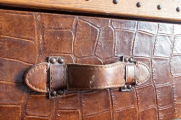A textured brown leather trunk with a leather strap buckle handle and metal studs along the wooden edge.