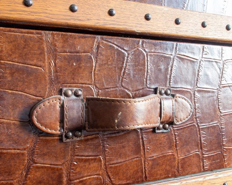 A textured brown leather trunk with a leather strap buckle handle and metal studs along the wooden edge.