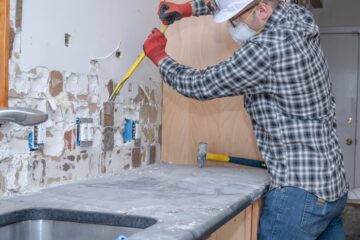 A man wearing gloves, a hard hat, safety glasses, and a face mask pries tile backsplash off a kitchen wall.