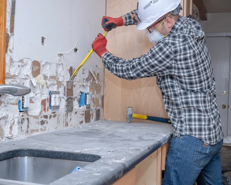A man wearing gloves, a hard hat, safety glasses, and a face mask pries tile backsplash off a kitchen wall.