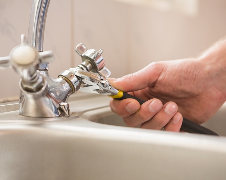 A hand holding pliers adjusts a chrome sink tap beside a white basin, tiled wall, and curved faucet.