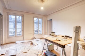 A room under renovation with woodworking tools, a workbench, and construction materials on a hardwood floor near tall windows.
