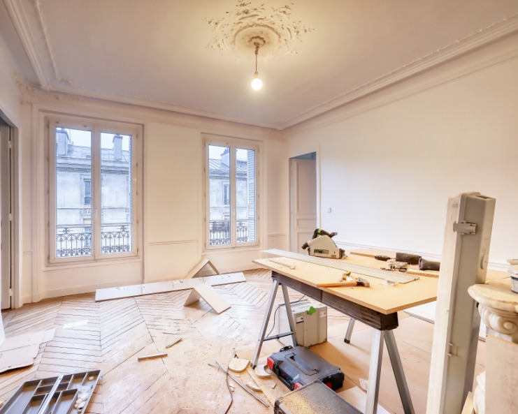 A room under renovation with woodworking tools, a workbench, and construction materials on a hardwood floor near tall windows.