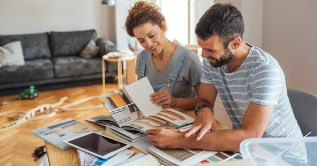 A couple compares home design catalogs and material samples at a table while planning a renovation in a bright living room.