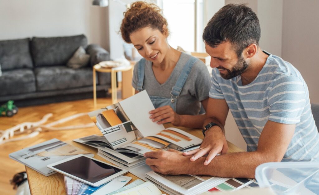 A couple compares home design catalogs and material samples at a table while planning a renovation in a bright living room.