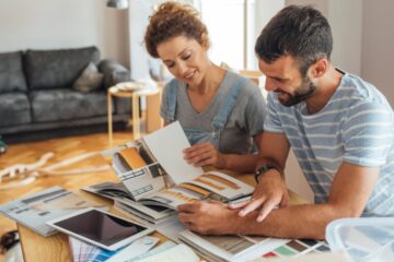 A couple compares home design catalogs and material samples at a table while planning a renovation in a bright living room.