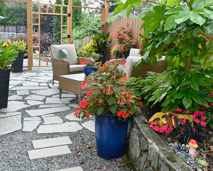A colorful backyard patio with stone pavers, outdoor seating, flowering plants, and a decorative wooden garden gate.