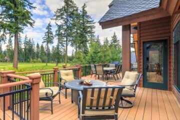 A wooden deck beside a cabin showing cushioned chairs around a fire table, with trees and open grass nearby.