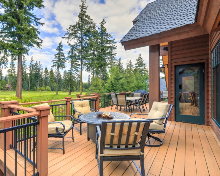 A wooden deck beside a cabin showing cushioned chairs around a fire table, with trees and open grass nearby.