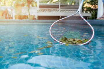 A person standing at a pool's edge, using a pool skimmer net to collect fallen leaves from blue water.