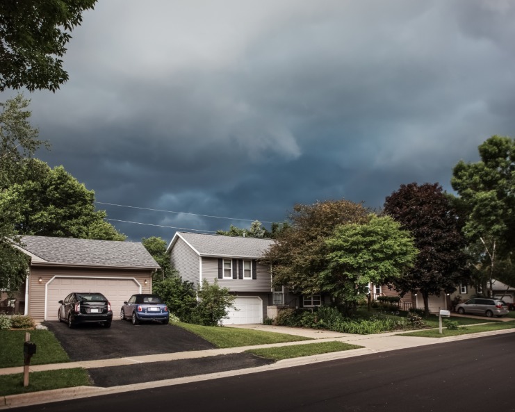 A suburban street with a row of houses and vehicles parked in front of them. Storm clouds are approaching the homes.