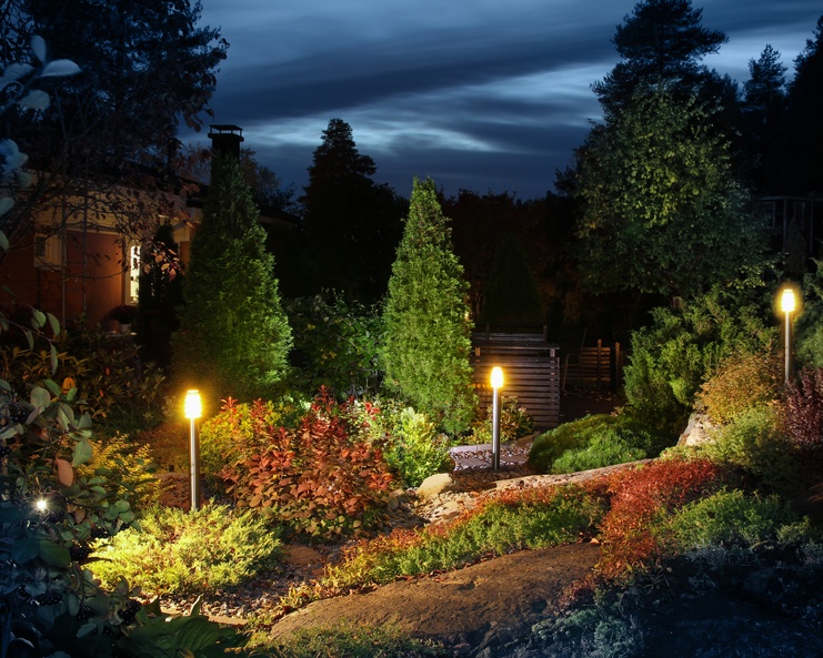 Warm garden lamps light a lush hillside of shrubs, small trees, and rocks beneath a deep blue evening sky.