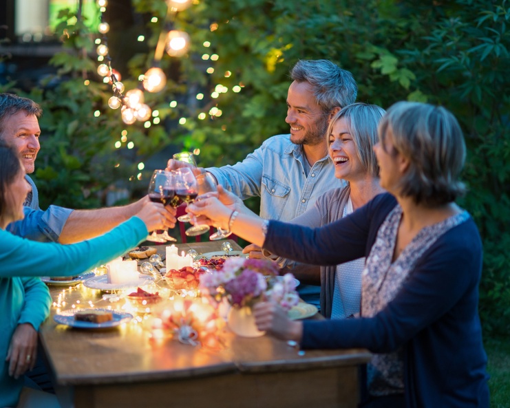 A group of adults seated outdoors at a table, raising glasses beneath glowing string lights at dusk.