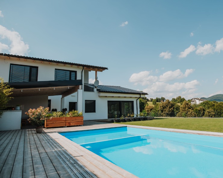 A modern white house beside a rectangular swimming pool, with a wooden deck, green lawn, and trees in the distance.