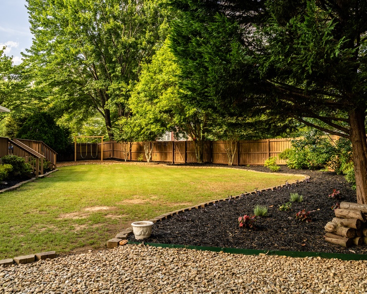 A residential backyard fully fenced in. There are some stronger trees along the fence and landscaping nearby.