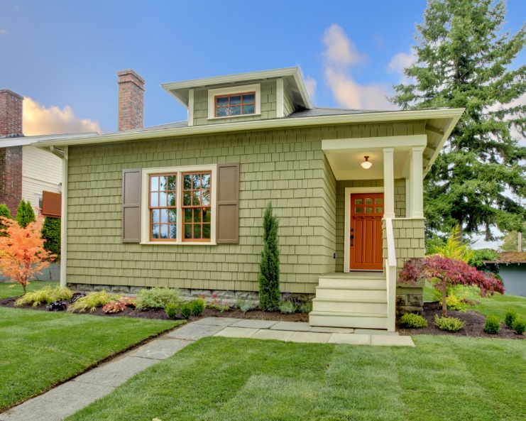 Landscaping surrounds a green Craftsman-style house that features brown shutters, white trim, and a covered doorway.